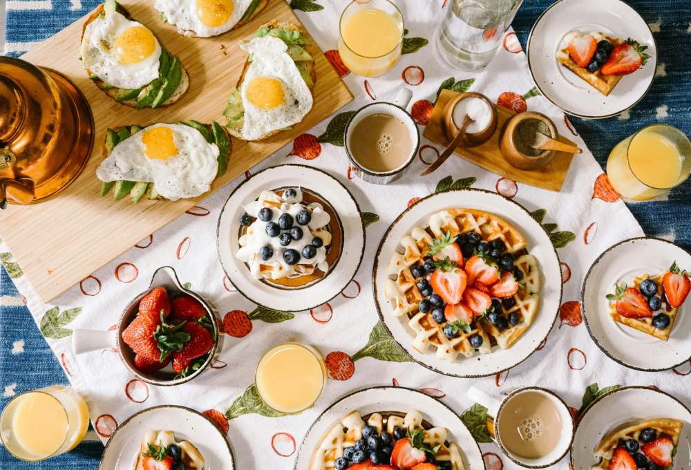 Desayuno con waffles, frutas, tostadas con huevo, café y jugo sobre mantel estampado con frutas.
