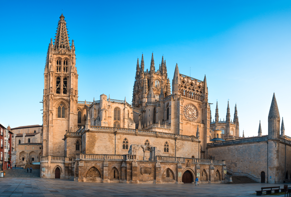 Catedral gótica con torres y rosetón en una plaza amplia, bajo un cielo despejado y azul.