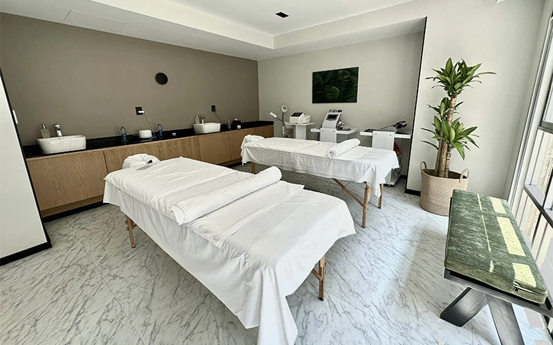 Sala de masajes moderna con dos camillas, decorada en tonos neutros y plantas. Luz natural entra por las ventanas.