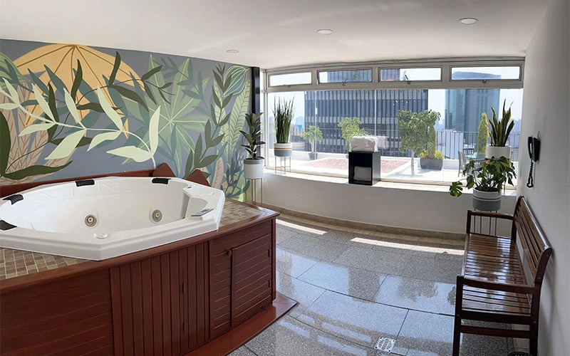 Jacuzzi en habitación luminosa con mural de plantas, vista a la ciudad y varias plantas en macetas junto a una ventana.
