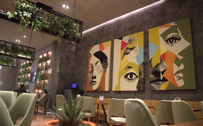 Cafetería moderna con paredes decoradas con arte colorido y plantas colgantes. Mesas y sillas en el interior.