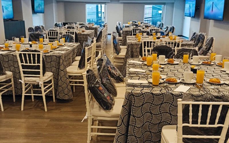 Salón con mesas decoradas, copas de jugo, tazas y sillas blancas, listo para un evento o banquete.