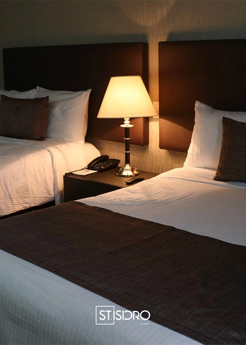 Dos camas en habitación de hotel con lámpara encendida sobre mesa de noche. Decoración en tonos marrones y blancos.