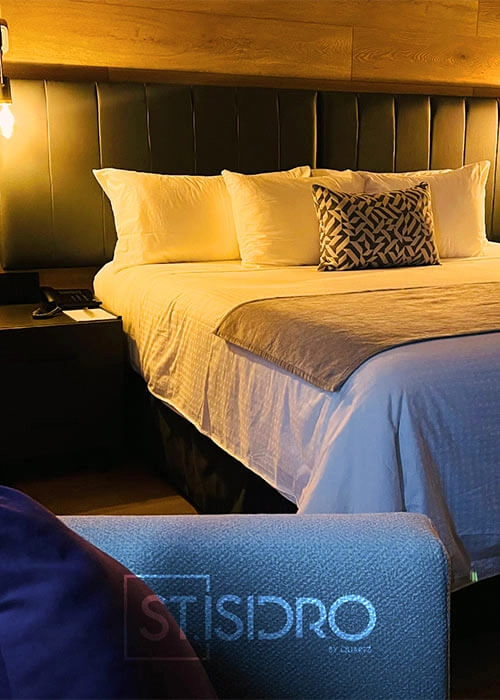 Cama de hotel acogedora con almohadas blancas y cojín decorativo. Mesa de noche a la izquierda y cabecera tapizada.