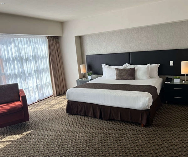 Habitación de hotel con cama grande, dos lámparas, sillón rojo y ventana con cortinas claras.