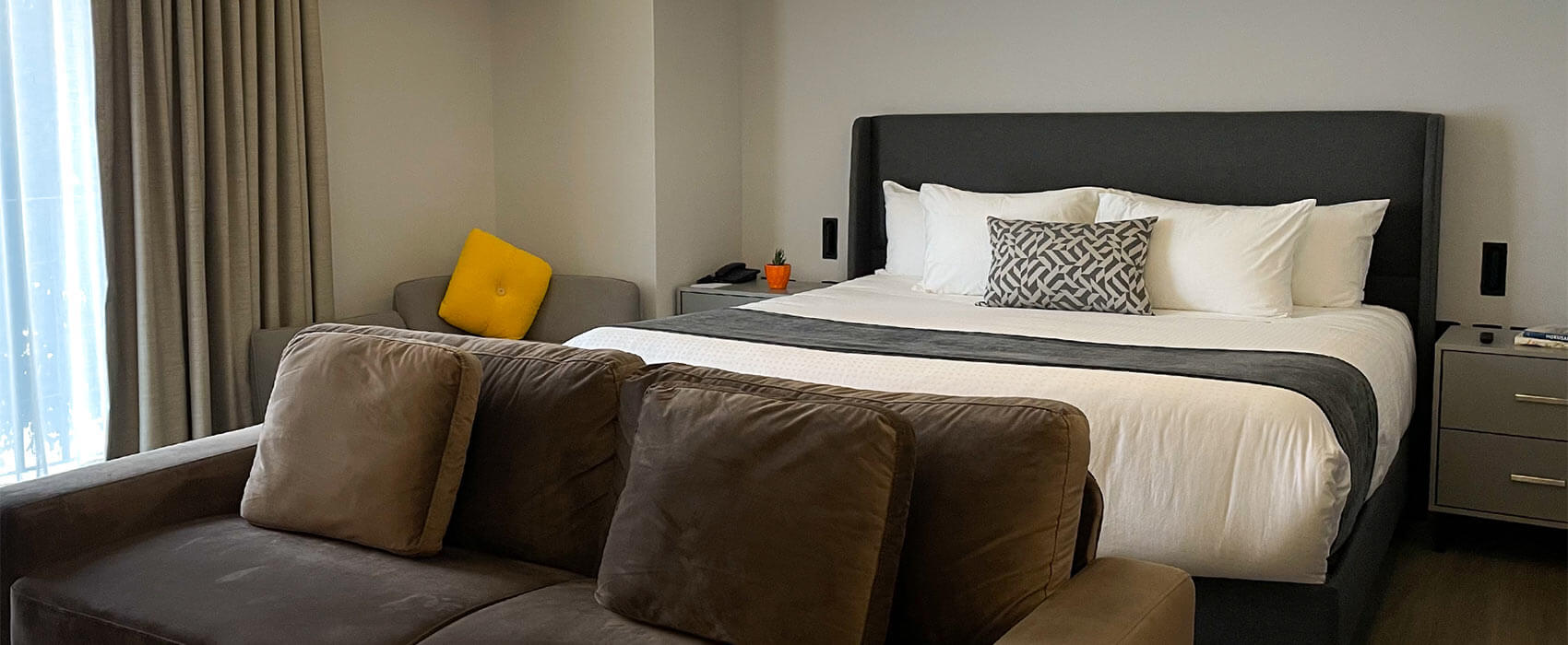 Habitación moderna con cama grande, sofá oscuro y decoración sencilla, cojín amarillo destacado en un sillón.