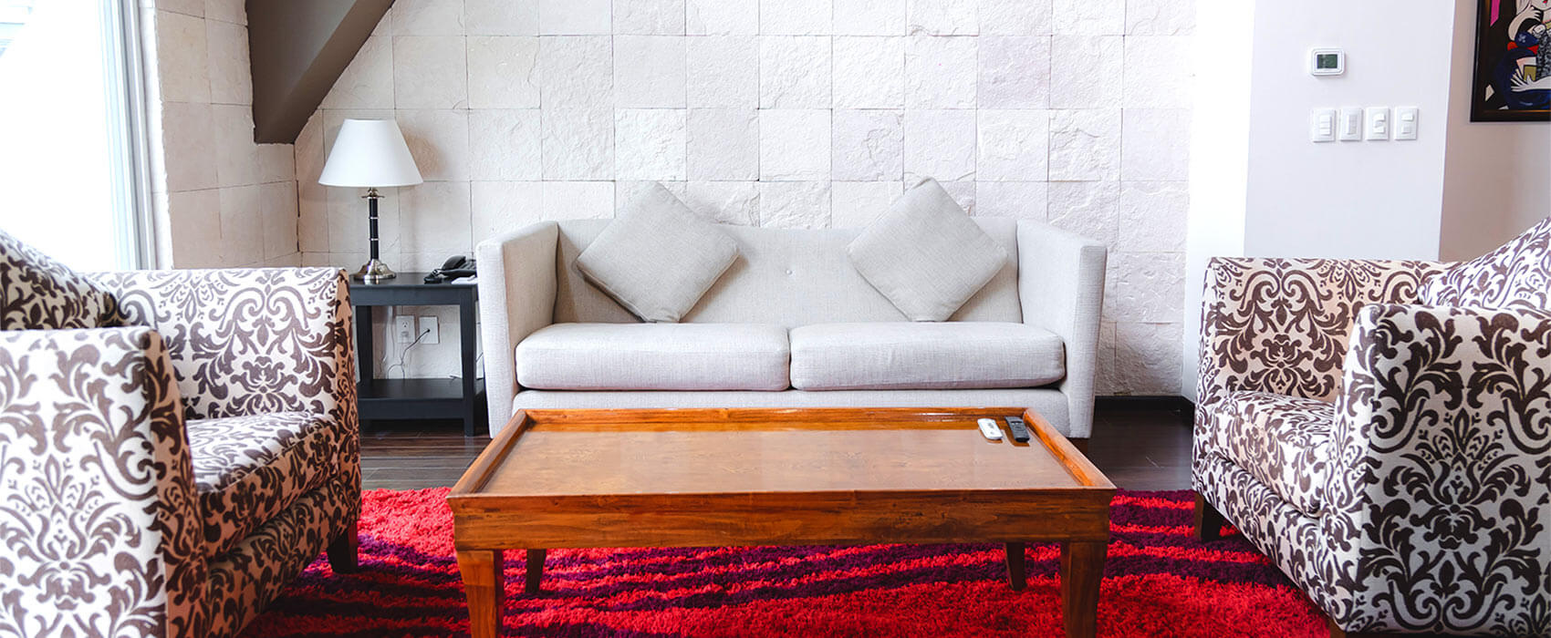 Sala con sofá gris, sillones estampados, mesa de madera, alfombra roja y lámpara sobre mesa lateral.