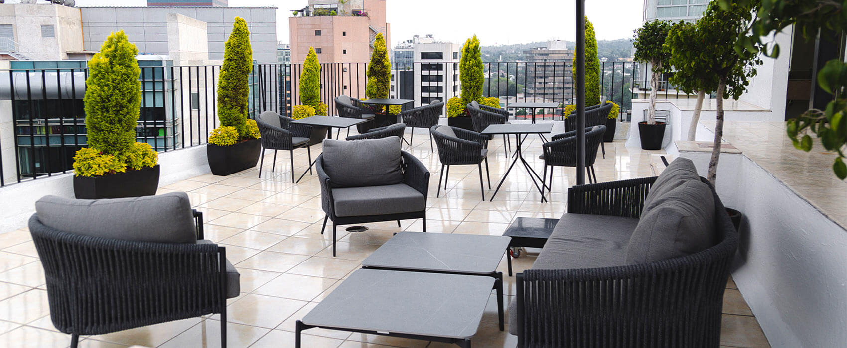 Terraza moderna con muebles oscuros, mesas, sillas y plantas en macetas. Vista urbana al fondo.