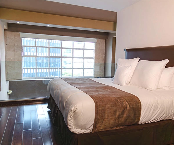 Habitación con cama grande, sábanas blancas, ventana amplia y piso de madera oscura iluminado por luz natural.
