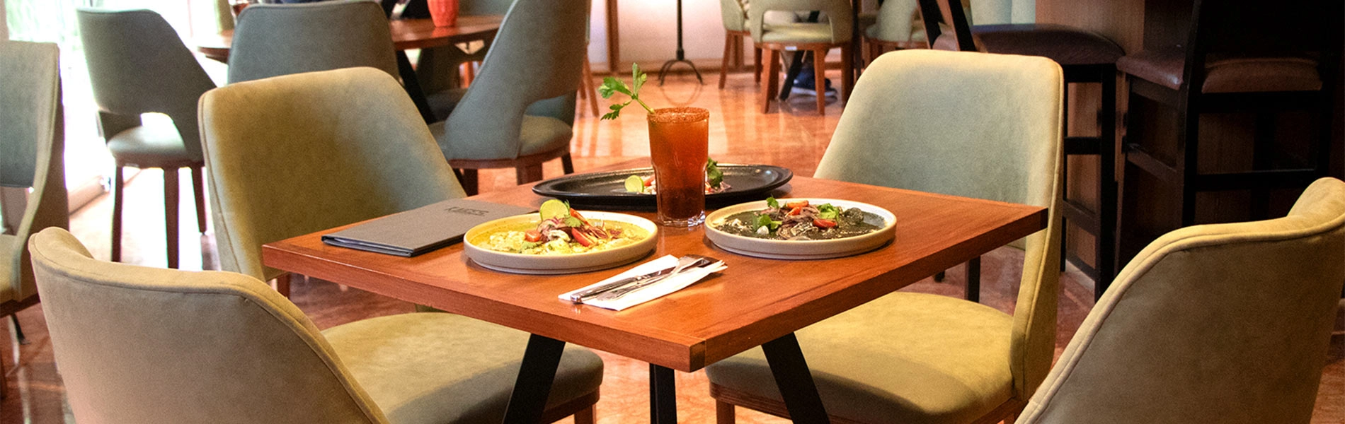 Mesa en un restaurante con platos de comida, bebida, servilletas, cubiertos y menú. Sillas acolchadas alrededor.