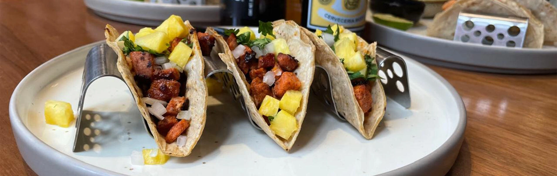 Tacos al pastor con piña y cilantro en soportes metálicos sobre un plato blanco, listos para disfrutar.