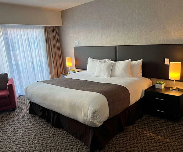 Habitación de hotel con cama grande, dos lámparas encendidas y ventana amplia con cortinas semi abiertas.