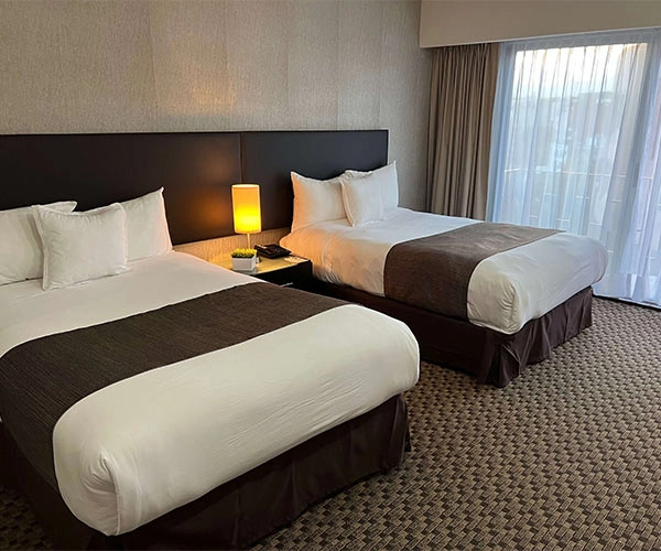 Habitación de hotel con dos camas dobles, lámpara de noche entre ellas y ventana con cortinas a la derecha.