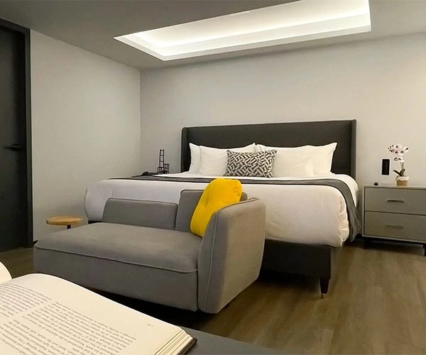Habitación moderna con cama grande, sofá gris con cojín amarillo, luz natural cenital y mesa de noche con decoraciones.