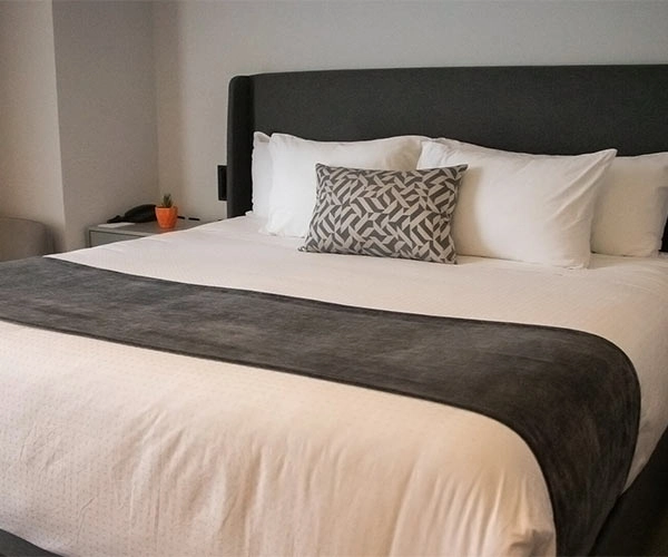 Cama de matrimonio con cabecero gris oscuro, cojines blancos, una almohada decorativa y manta gris en habitación minimalista.