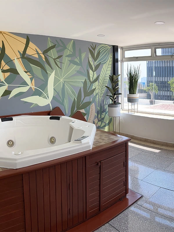 Jacuzzi interior frente a un mural con hojas verdes y amarillas; plantas junto a una ventana soleada.
