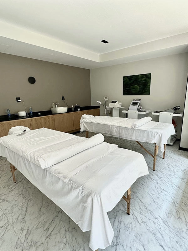 Sala de masajes moderna con dos camillas cubiertas de sábanas blancas, equipo de spa y decoración sencilla.