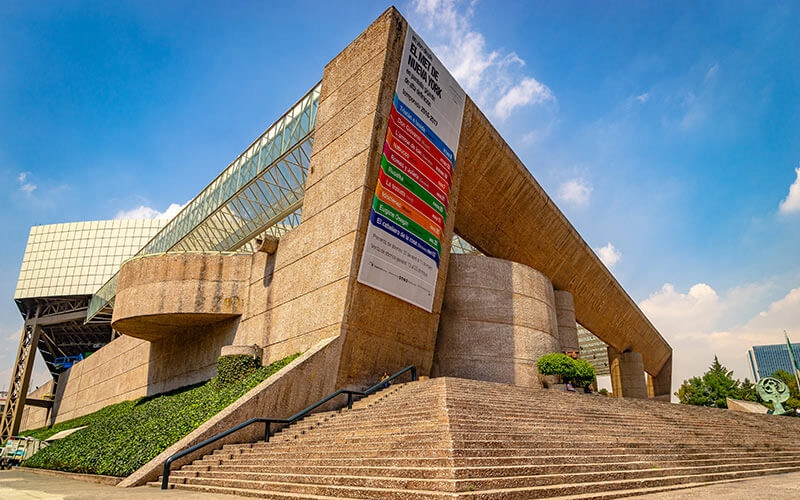 Edificio moderno con fachada inclinada, escalera amplia y un cartel colorido. Cielo despejado de fondo.