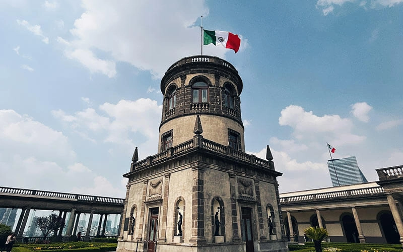 Castillo de Chapultepec con una bandera de México ondeando en lo alto, cielo azul y algunas nubes de fondo.