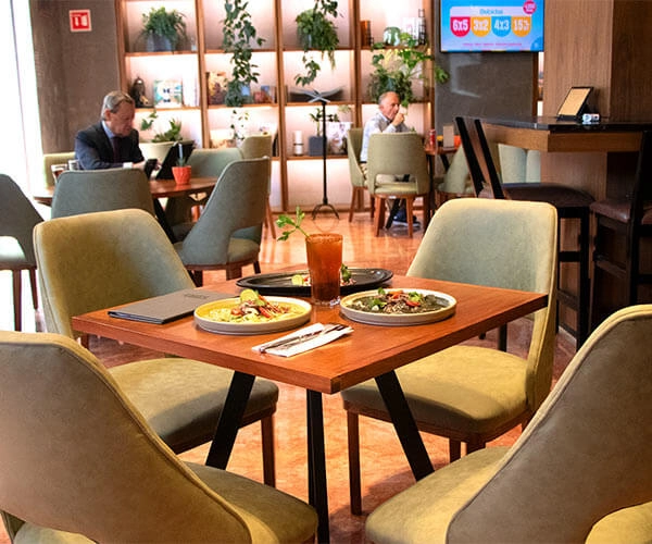 Mesa en restaurante con comida y bebida. Al fondo, personas sentadas y estantería con plantas decorativas.