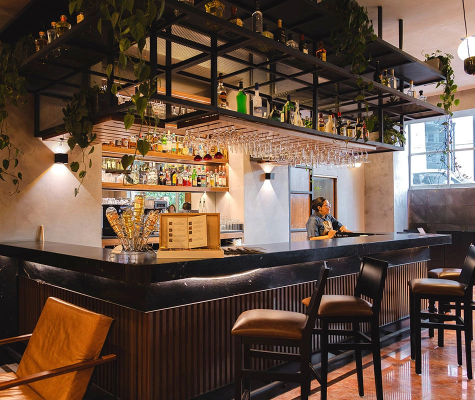 Bar moderno con estanterías llenas de botellas, copas colgantes y un barman detrás de una barra oscura.