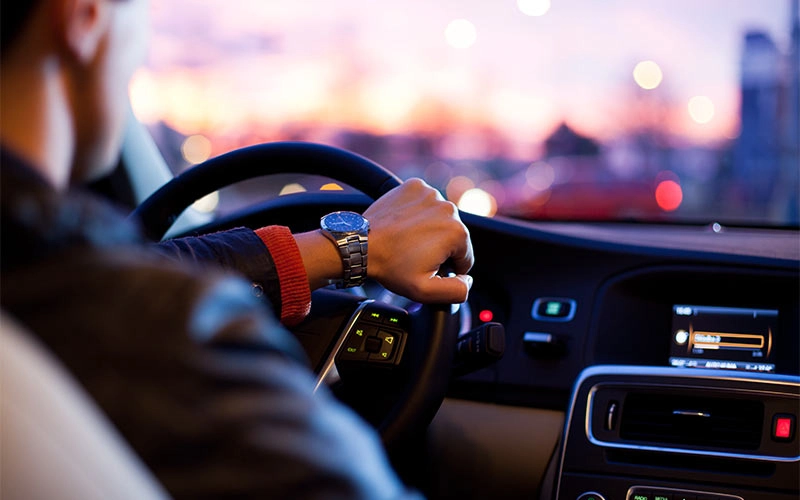 Persona conduciendo un coche al atardecer, con las manos en el volante y luces de la ciudad en el fondo.