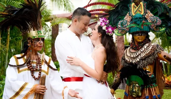 Pareja abrazada con trajes blancos rodeada por personas con coloridos trajes tradicionales indígenas en un entorno tropical.