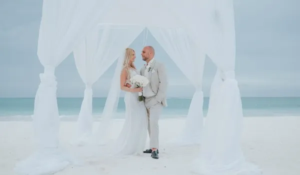 Pareja de novios bajo un dosel blanco en una playa, con el mar de fondo y cielo nublado.