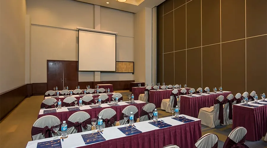 Sala de conferencias con sillas y mesas arregladas, pantalla grande y agua embotellada.