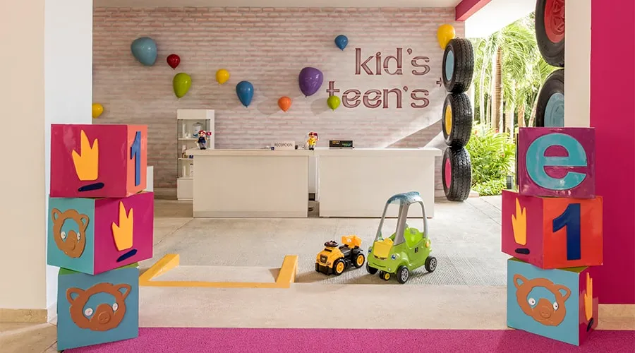 Área infantil con juguetes, bloques y decoración colorida; fondo con pared y globos.
