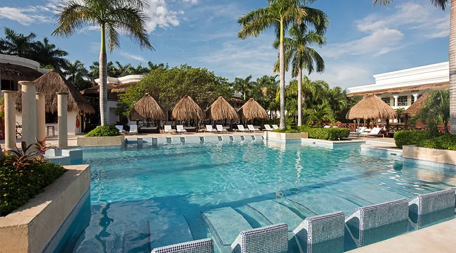 Piscina con palapas, palmeras y tumbonas, bajo un cielo azul.