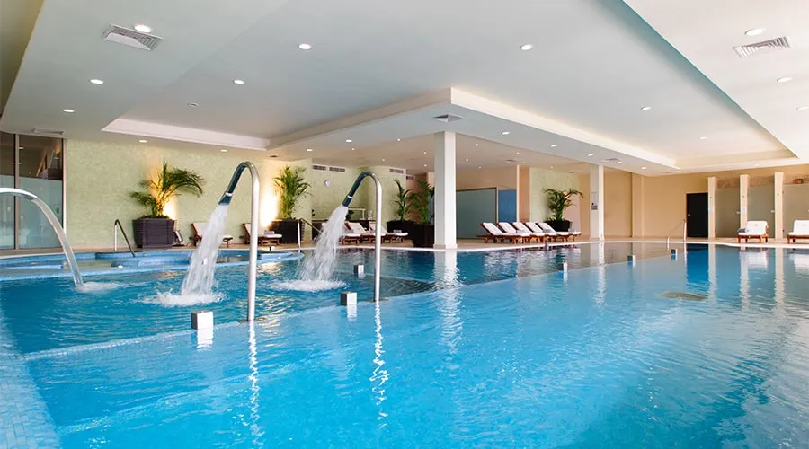 Piscina cubierta con chorros de agua, tumbonas y plantas decorativas alrededor.