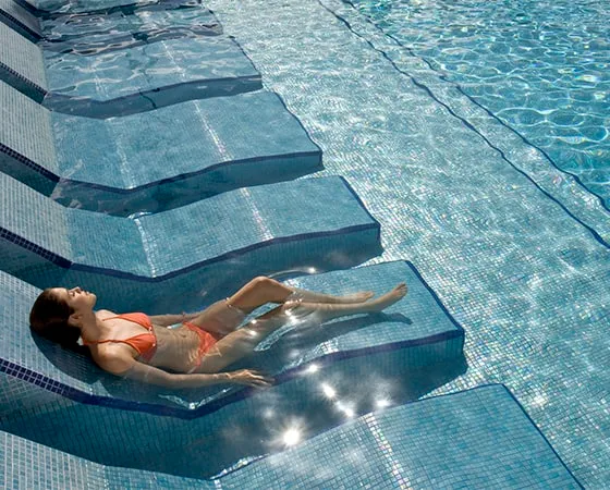 Persona descansando en una piscina sobre un sillón sumergido. Lleva traje de baño naranja.