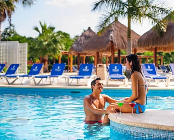 Pareja en una piscina disfrutando de un cóctel y el sol, rodeados de palmeras y tumbonas azules.