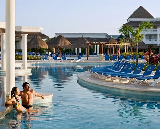 Pareja en la piscina de un resort tropical, rodeada de sombrillas y tumbonas azules bajo un cielo despejado.