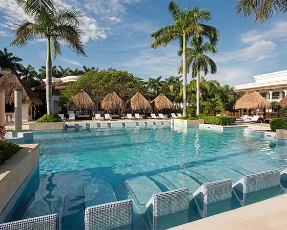 Piscina al aire libre rodeada de palmeras y palapas, con sillones sumergidos en el agua. Ambiente tropical y soleado.