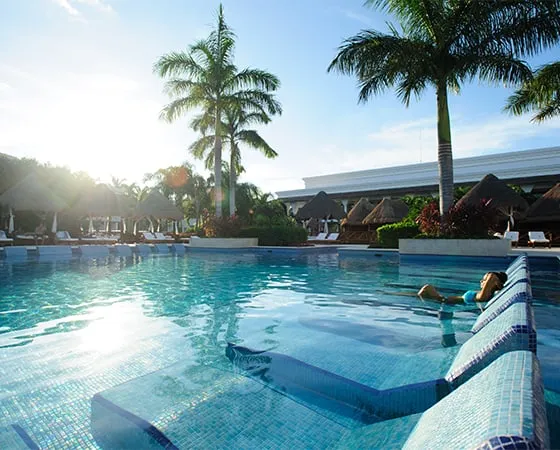 Persona relajándose en una piscina al aire libre, rodeada de palmeras y sombrillas bajo un cielo despejado.