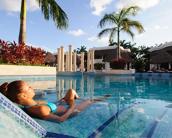 Mujer relajándose en una piscina bajo el sol, rodeada de palmeras y arquitectura tropical.