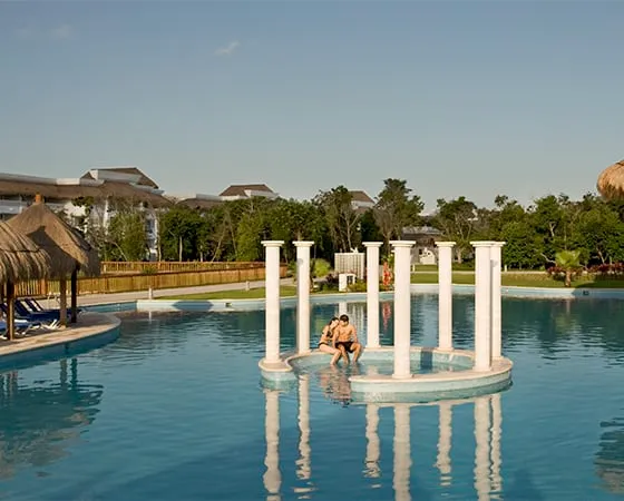 Persona sentada en una estructura circular con columnas en medio de una piscina, rodeada de vegetación y edificios.