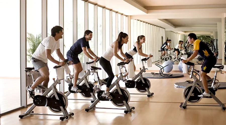 Grupo de personas haciendo spinning en un gimnasio con espejos y ventanas amplias.