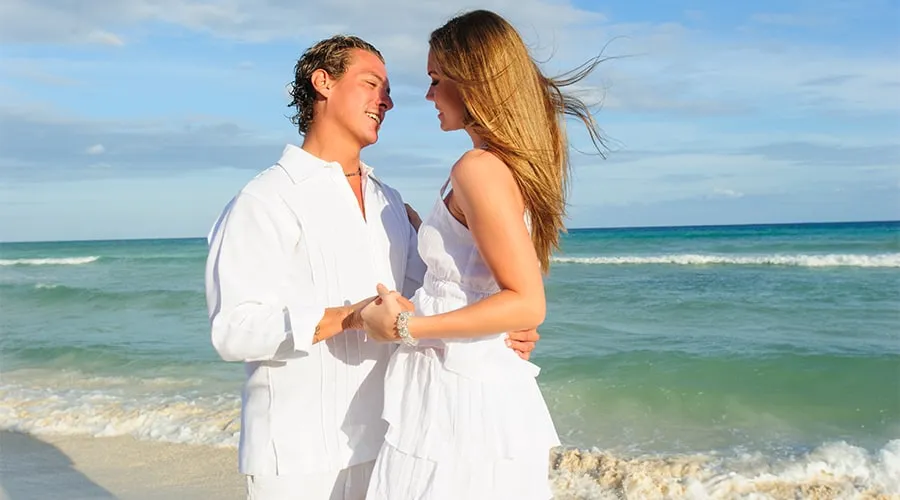 Pareja vestida de blanco se mira con afecto en la playa, con el mar y el cielo azul de fondo.