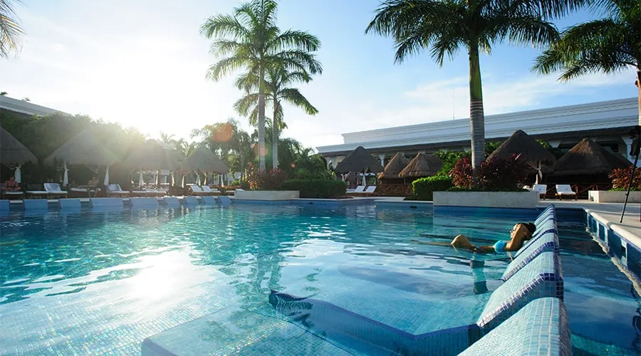 Piscina al aire libre con palmeras y cabañas al fondo, bajo un cálido sol, una persona relajándose en el agua.
