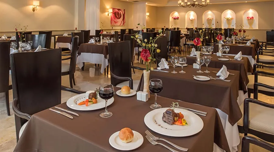 Restaurante elegante con mesas vestidas, platos de carne, copas de vino y decoración floral. Ambiente acogedor y sofisticado.