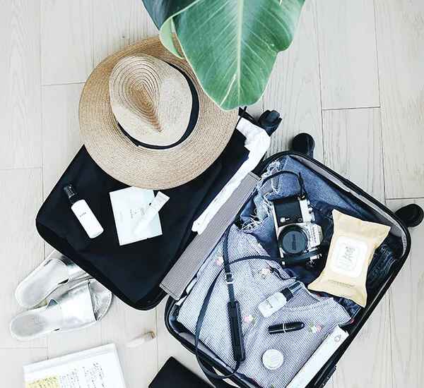 Maleta abierta con ropa, cámara, sombrero de paja, artículos de tocador y sandalias, lista para un viaje.