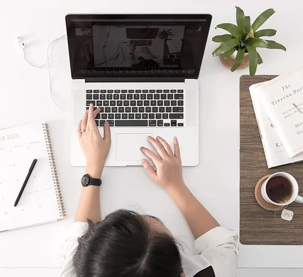 Persona usando laptop en escritorio con libreta, libro, taza de café y planta. Vista desde arriba.