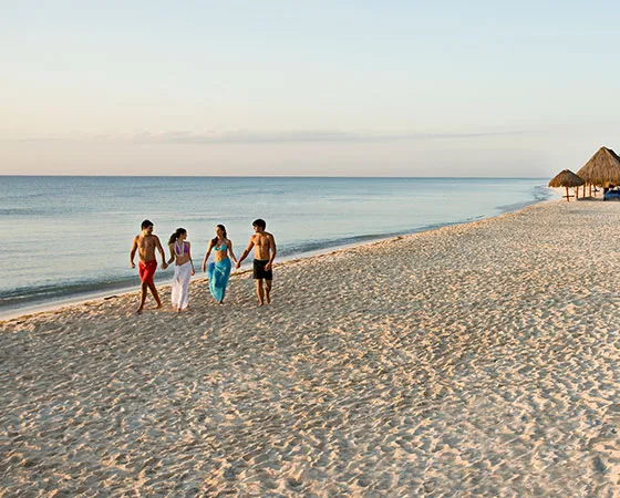 Cuatro personas caminando por la playa al atardecer, con sombrillas de paja en el fondo y el mar tranquilo a la izquierda.