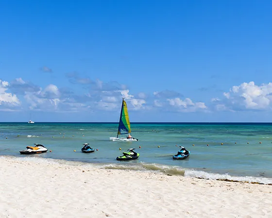 Playa con motos acuáticas y un velero en el mar bajo un cielo azul con nubes.