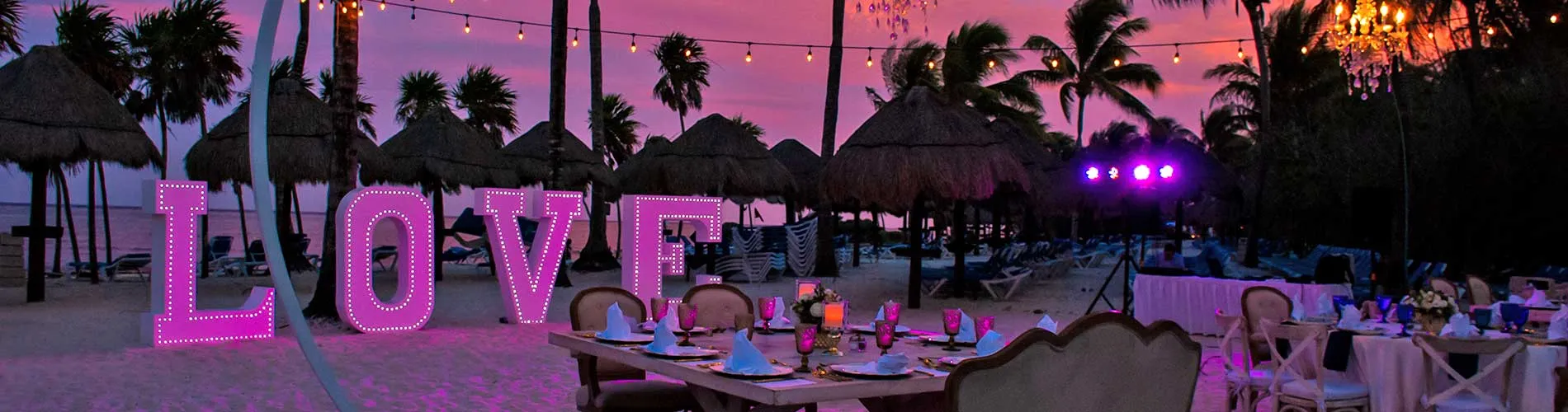 Playa al atardecer con luces de "LOVE" y mesas decoradas para un evento romántico.
