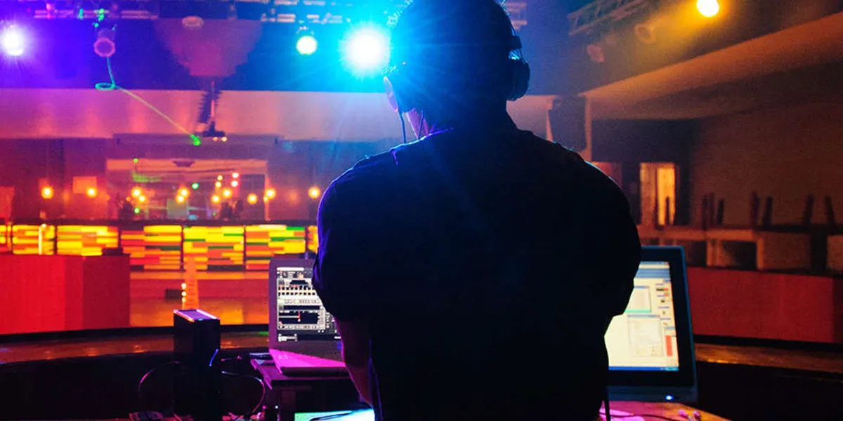 DJ en una cabina iluminada por luces de colores, con pantallas de computadora al frente en un club nocturno.