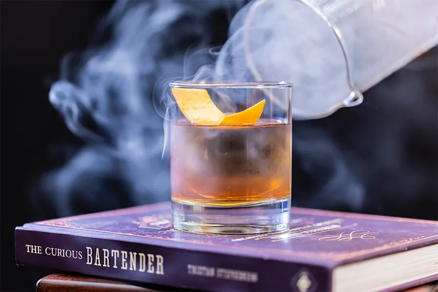 Vaso de cóctel con cáscara de naranja y humo, sobre un libro titulado "The Curious Bartender".